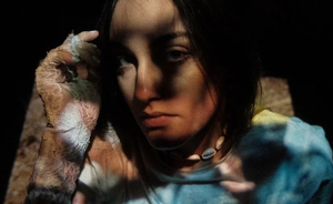 Bandfoto Mar Buss(Porträtaufnahme)

Eine Person sitzt im Halbdunkel und wird von Licht- und Schattenspielen überzogen. Das Gesicht ist nachdenklich und ernst, die Augen blicken leicht nach unten. Die Person trägt ein buntes, batikartiges Oberteil und eine Muschelkette. Die linke Hand ist hochgehoben und mit einem Verband oder Spitzenhandschuh bedeckt. Der Hintergrund ist dunkel und lässt die Person geheimnisvoll und introspektiv wirken.